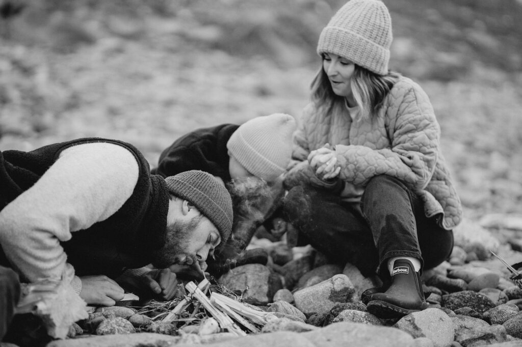 A family sitting around a small fire on the beach, toasting marshmallows as the sky turns soft and grey.