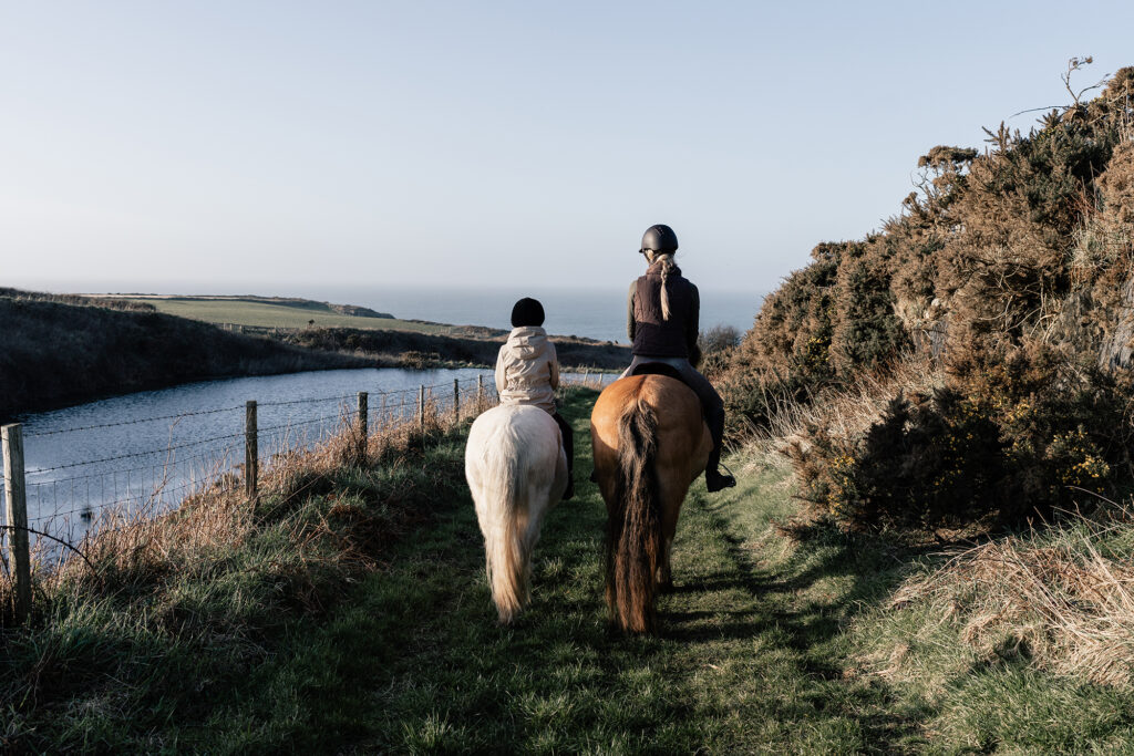 A natural family photoshoot in St Davids, Pembrokeshire, with a mother and daughter riding horses at sunset on the coast