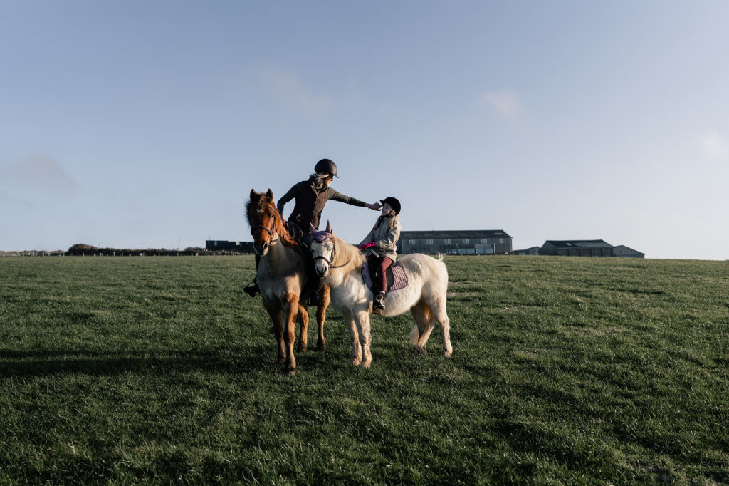 A natural family photoshoot in St Davids, Pembrokeshire, with a mother and daughter riding horses at sunset on the coast