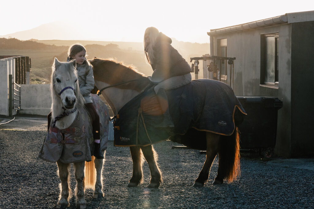 A natural family photoshoot in St Davids, Pembrokeshire, with a mother and daughter riding horses at sunset on the coast